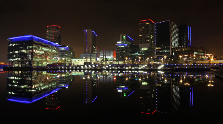 A night time photo of MediaCity, Salford, Manchester showing the neon lights reflecting against the water